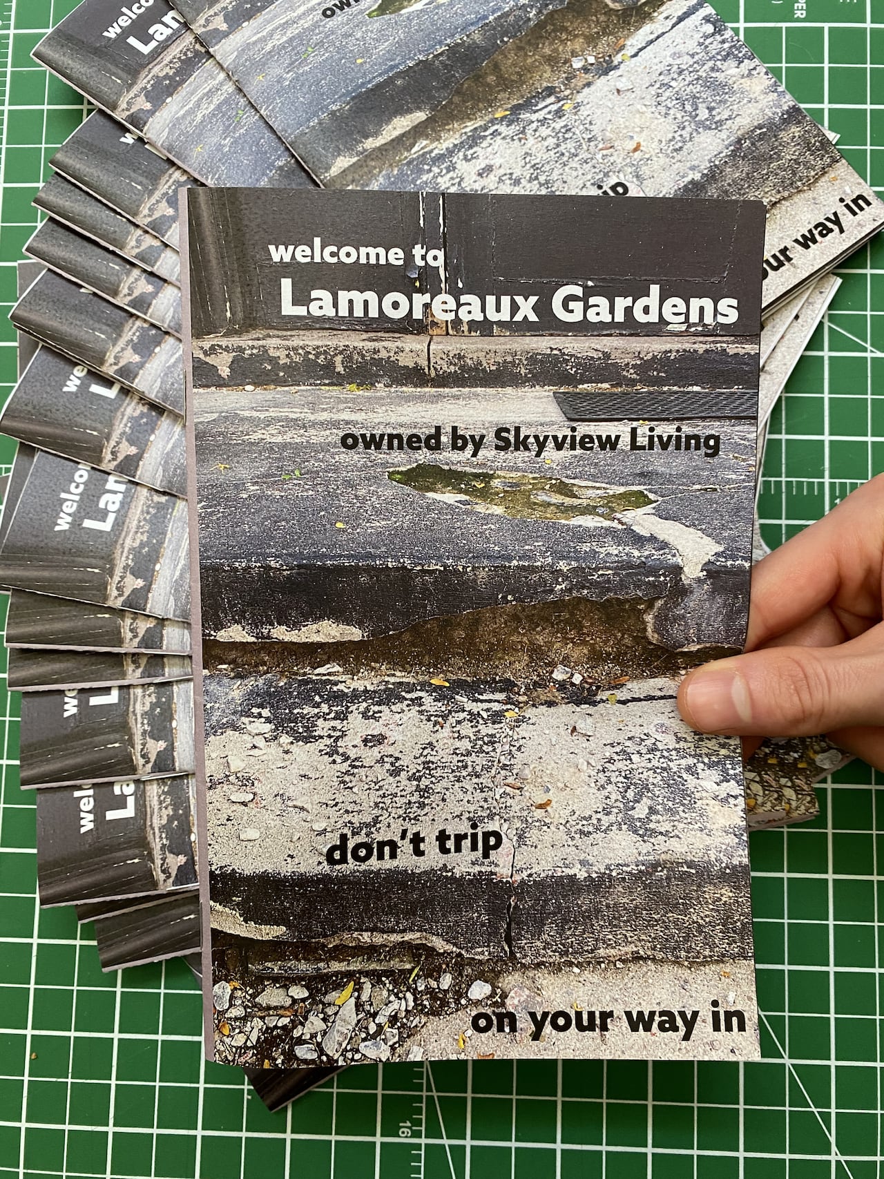 Over a green, gridded cutting mat, a hand holds a publication titled "Welcome to Lamoreaux Gardens." The cover shows a cracked cement stairway with the text "Don't trip on your way in." More than a dozen copies are fanned out underneath.