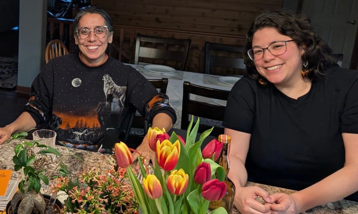 Two women smile next to a bouquet around a dinner table.