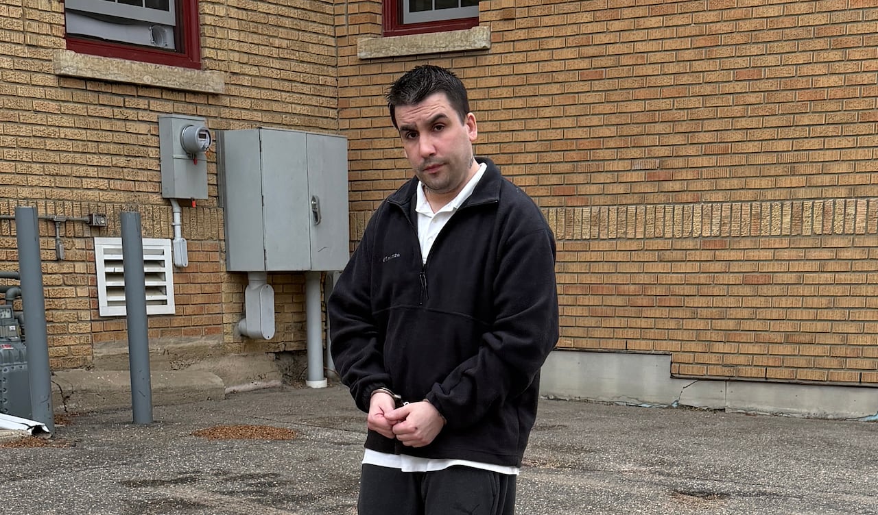 A man wearing black and in handcuffs walks behind a building.
