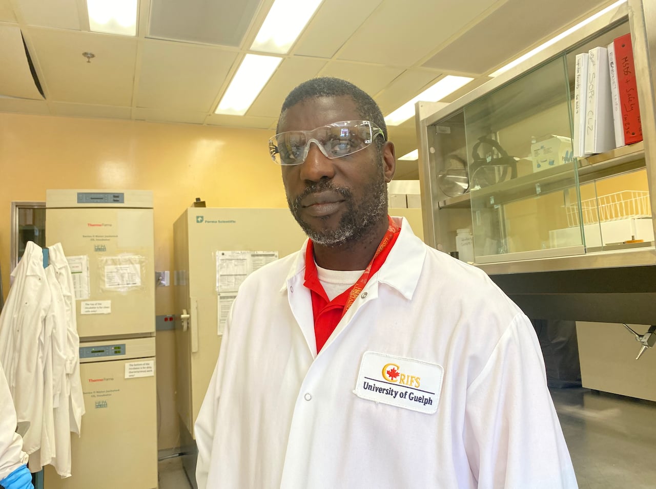A man stands wearing goggles, red shirt and lab coat with an embroidered label on the chest reading RIFS and University of Guelph under it.