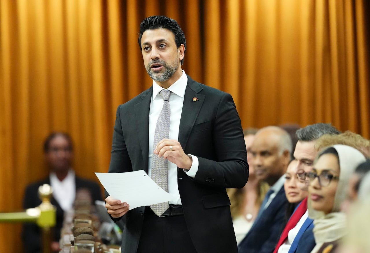 A man in a suit holds a piece of paper as he speaks inside a legislative chamber.
