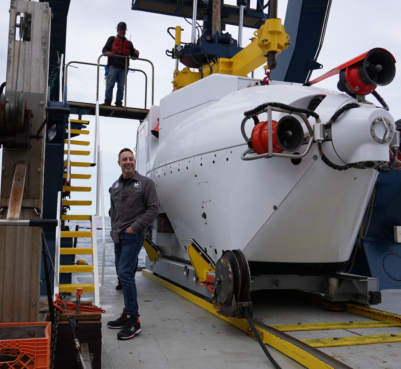 A man smiles as he stands beside a large, white submersible vehicle. Another man wearing a safety helmet stands on a platform above him.