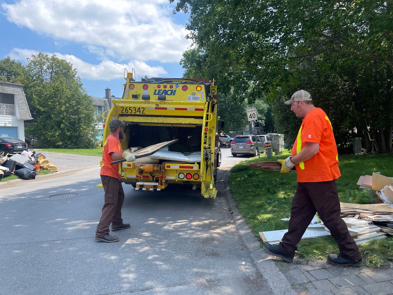 Two waste workers move flood-damaged wood into a garbage truck on a residential street.