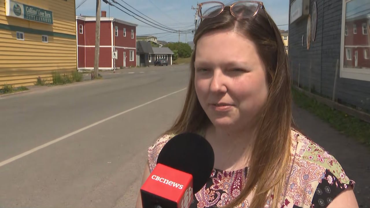 A woman speaks into a CBC microphone while standing outside. A street is visible to her right and the entrance to a one-storey building to her left.