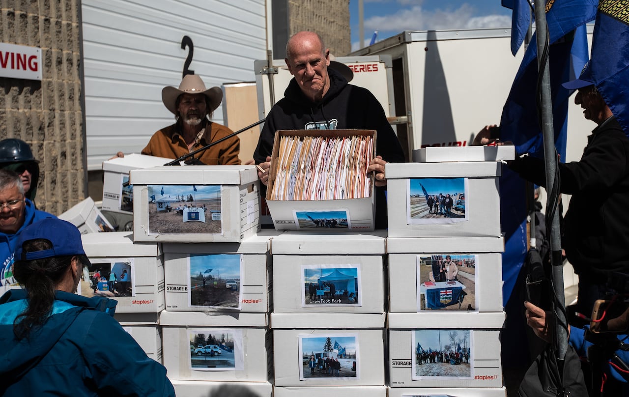 A man displays a box filled with paper, while standing behind a stack of multiple other boxes.