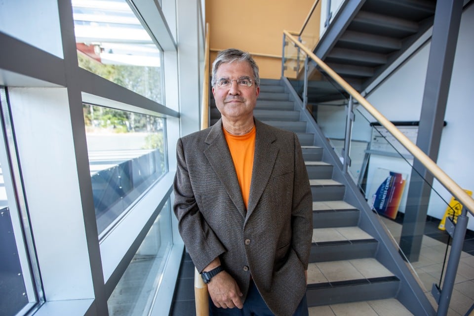 A smiling man with glasses, wearing a brown blazer over an orange shirt, stands next to a staircase and large windows