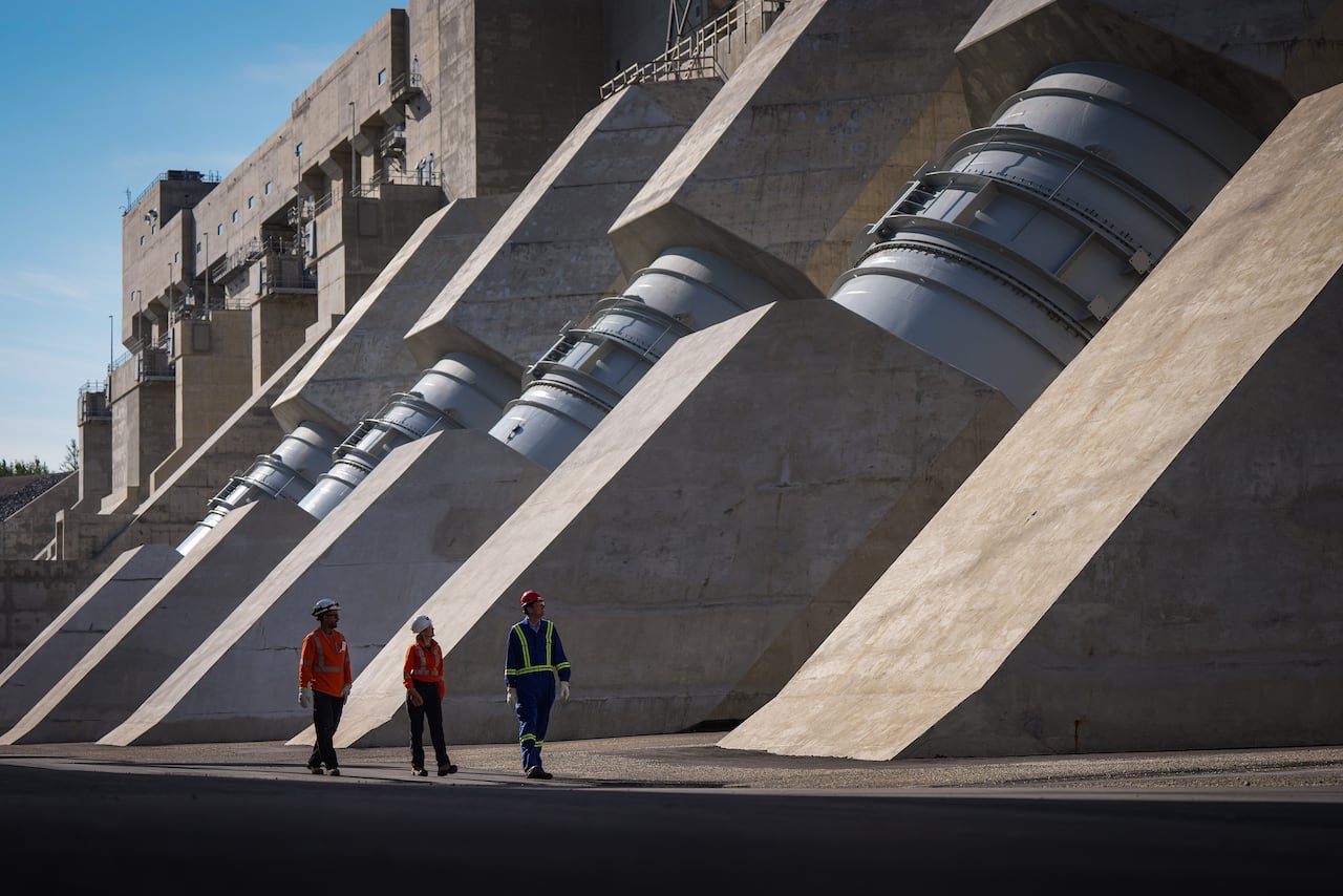 Three people in safety equipment walk in front of a massive concrete dam with huge, cylindrical tunnel components visible on the outside.
