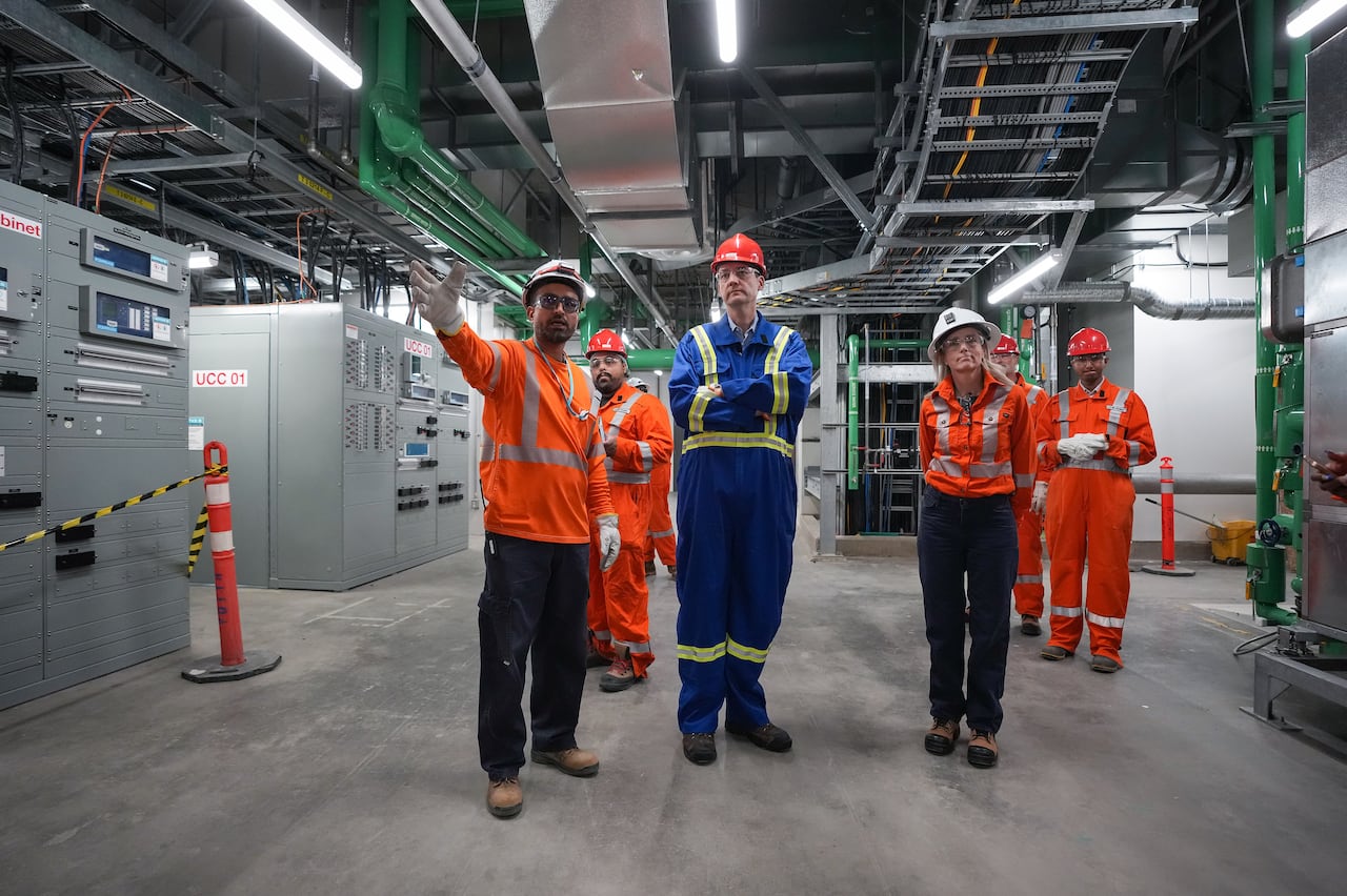 A group of people wearing safety equipment stand in a brightly lit room with a lot of ducts and control equipment.