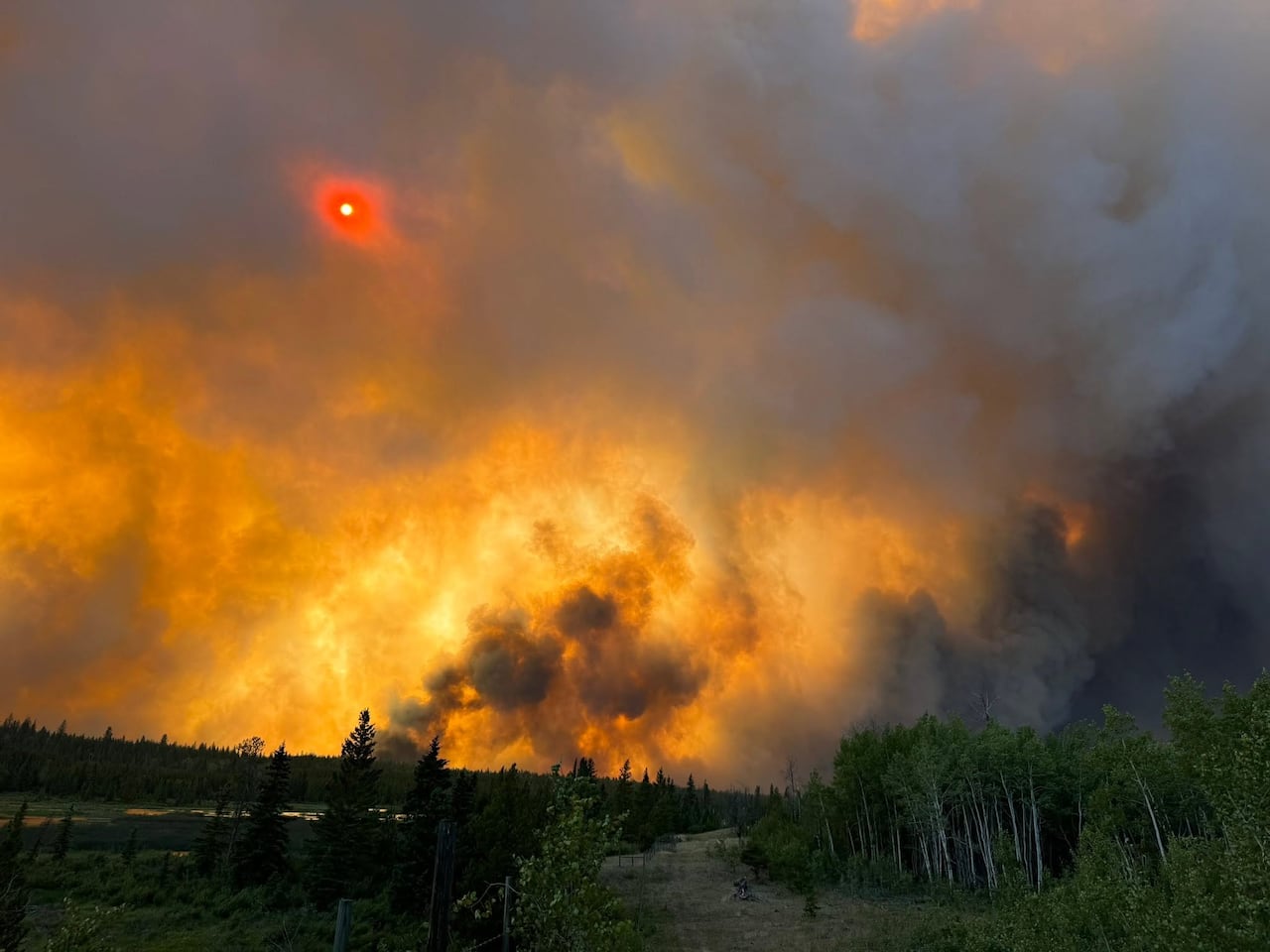 A massive wall of orange-lit smoke rises above a forested hillside as the sun glows red through the haze.
