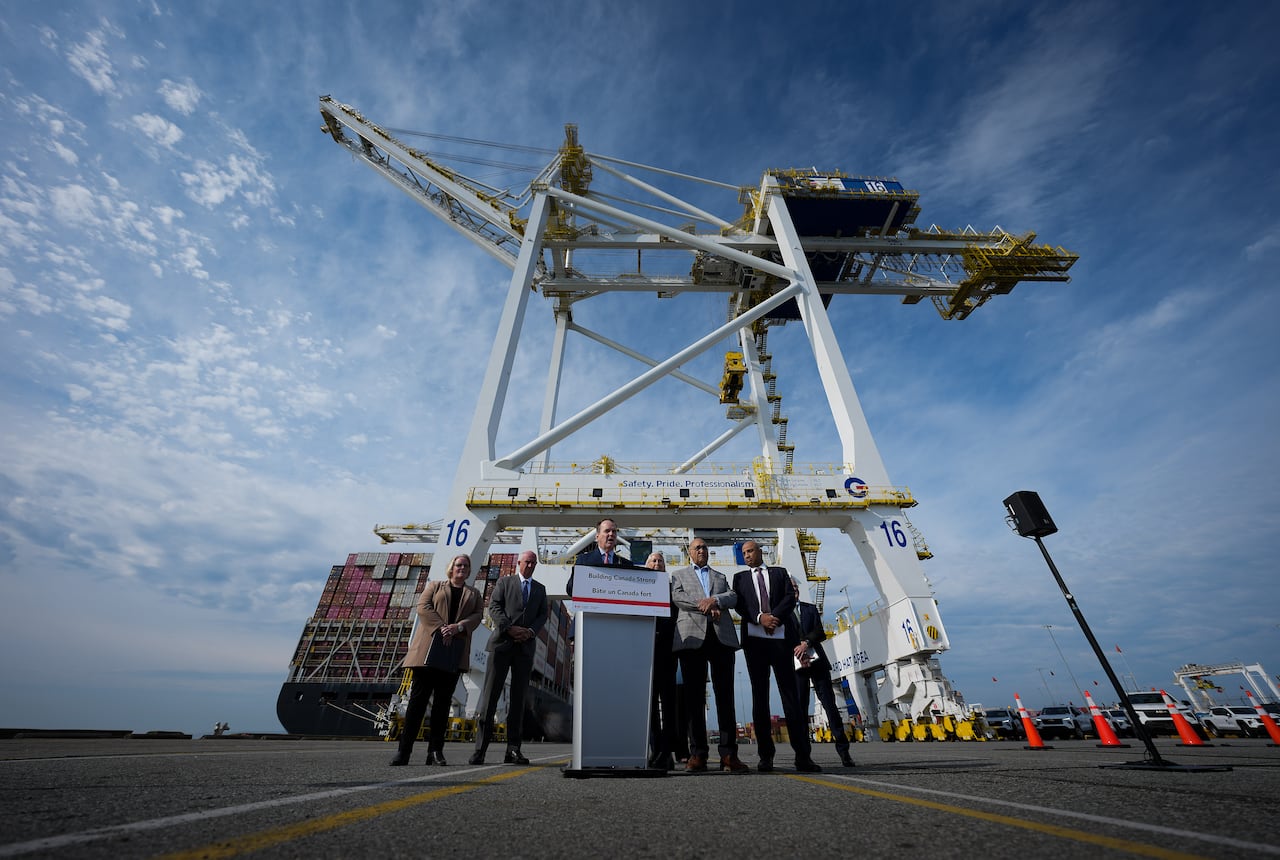 A group of people stand on a stretch of pavement under a large crane as a container ship is unloaded behind them.