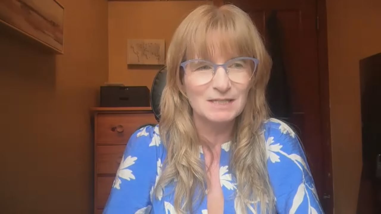 A woman with long blonde hair and bangs wearing glasses and a blue shirt, speaks to the camera.
