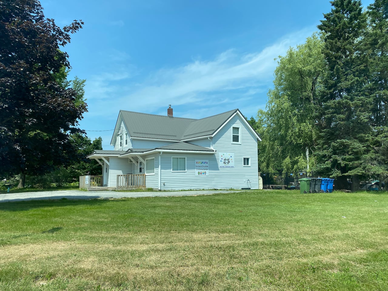 A two-storey building with grey/blue vinyl siding is pictured under a blue sky surrounded by trees and a freshly-mowed lawn.