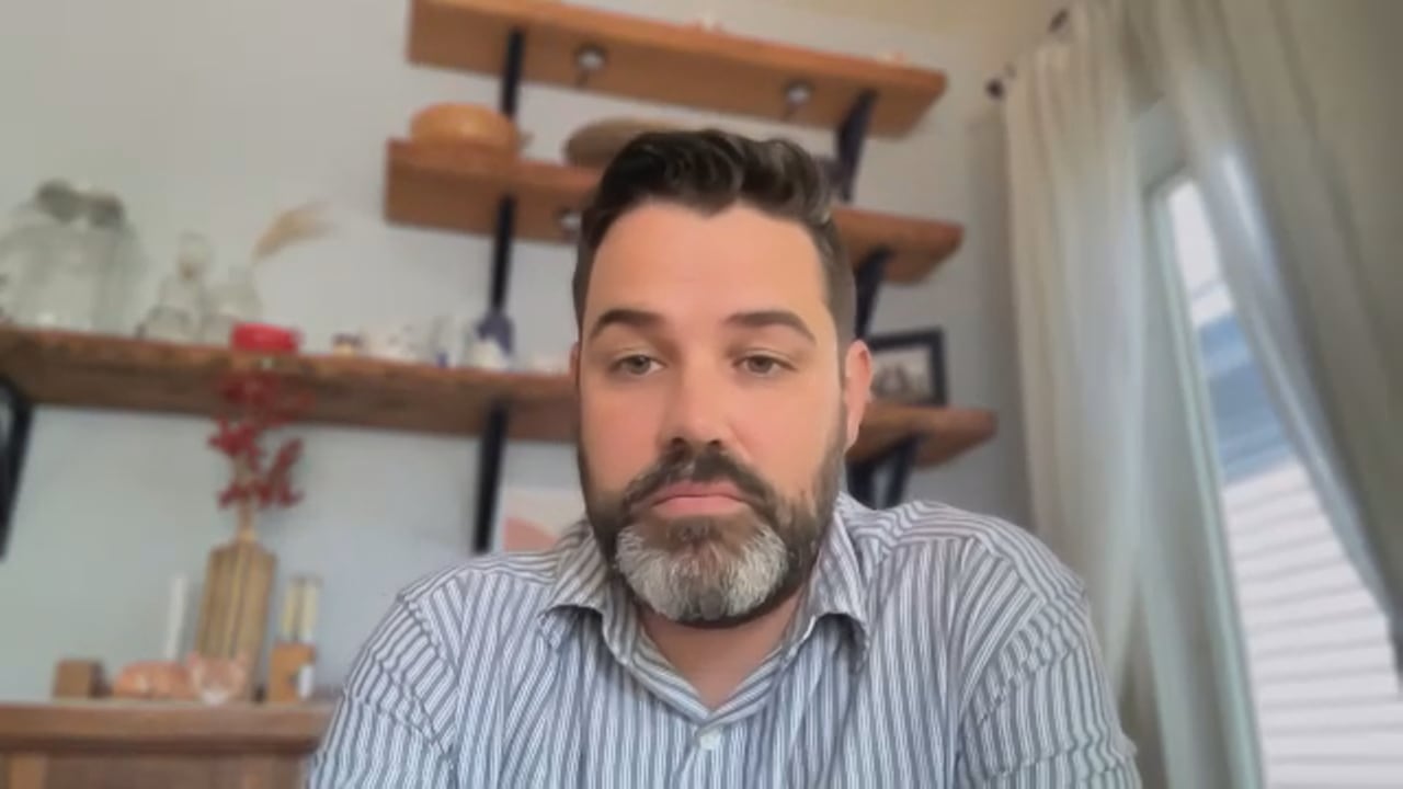 A man with brown hair, a mustache and a greying beard looks towards the camera. He is wearing a blue and white striped dress shirt and appears to be sitting at home.