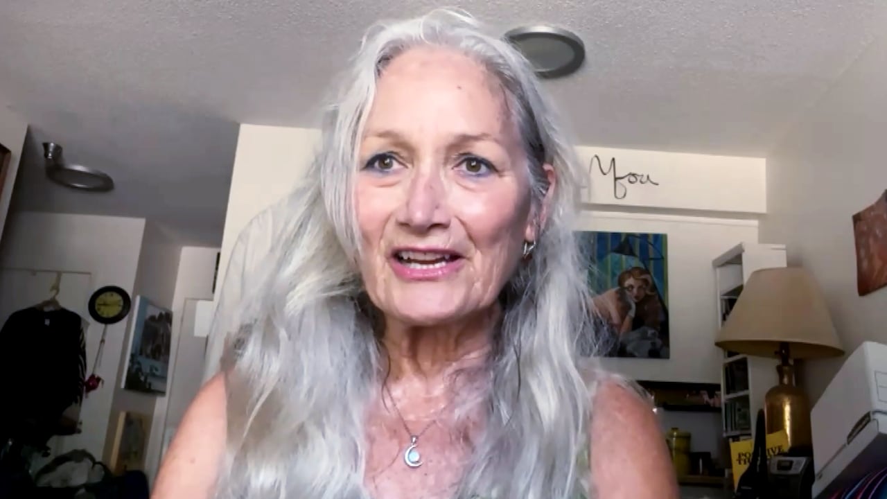 A woman with long grey hair in a sundress is pictured looking at the camera from the shoulders up.