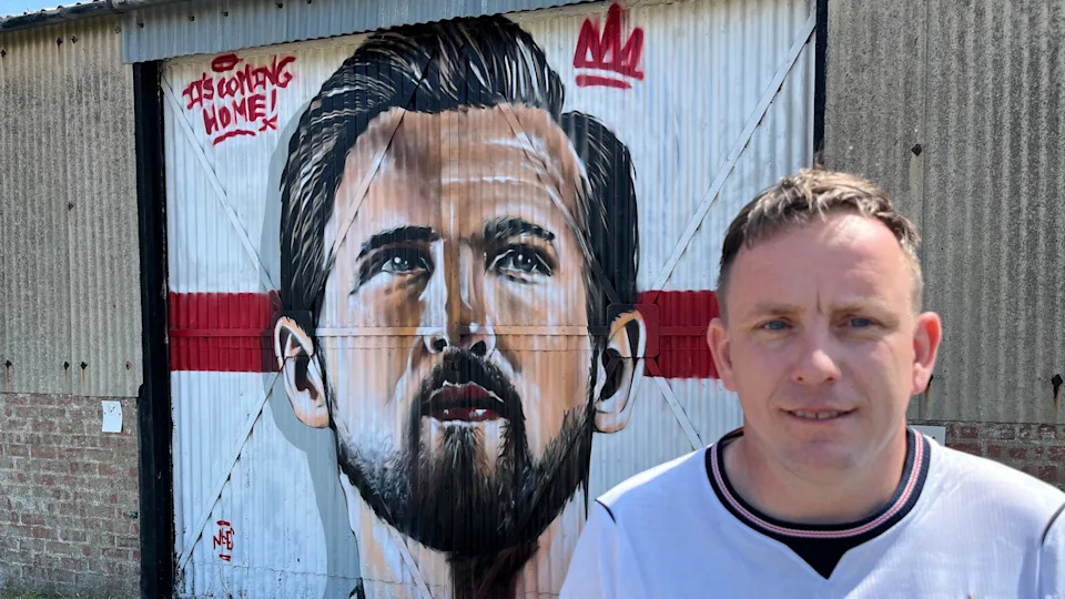 Street artist Nathan Murdoch looking at the camera. His mural of Harry Kane is behind him on a building.