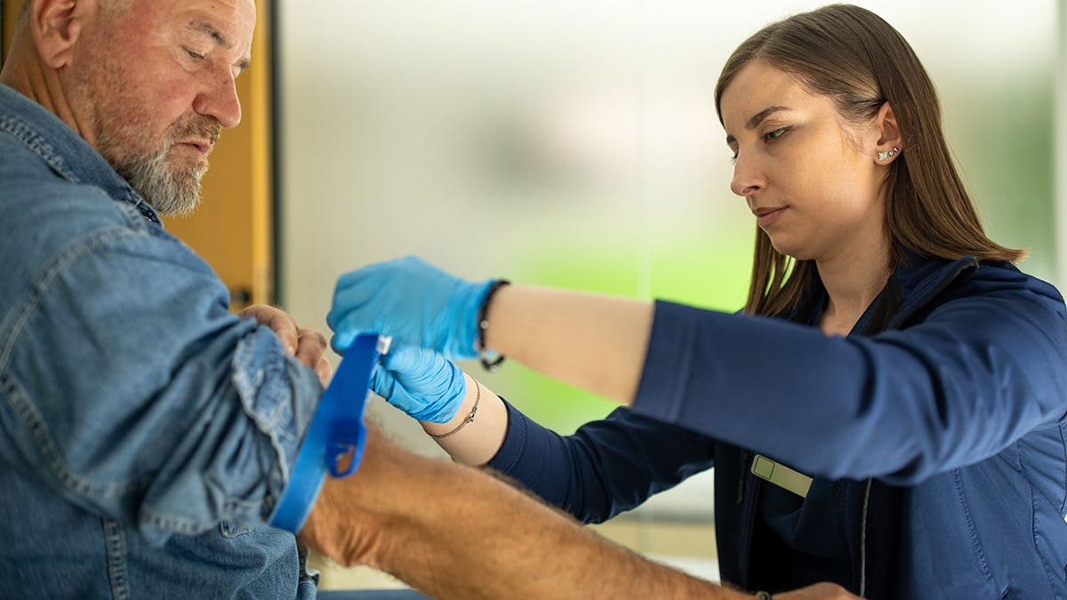 Female doctor prepares senior man's arm for blood test in clinic examination room.
