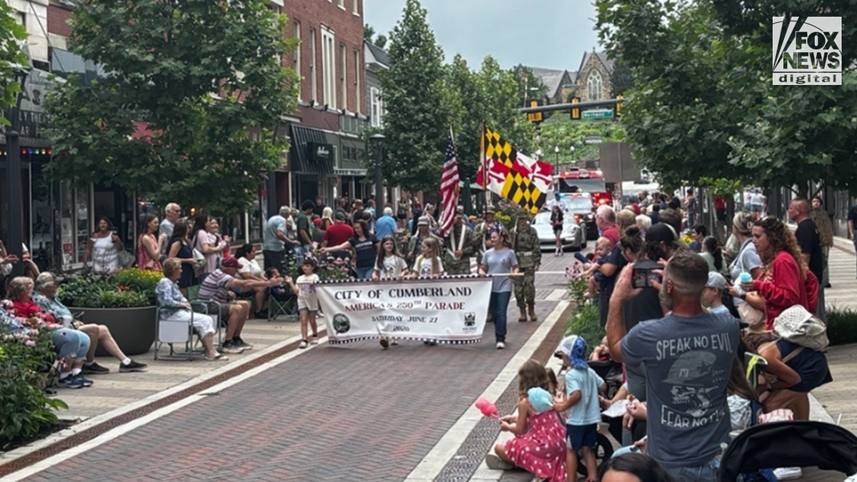 Parade participants carry a City of Cumberland America 250 banner and American and Maryland flags through downtown Cumberland, Maryland, as spectators line both sides of the street.
