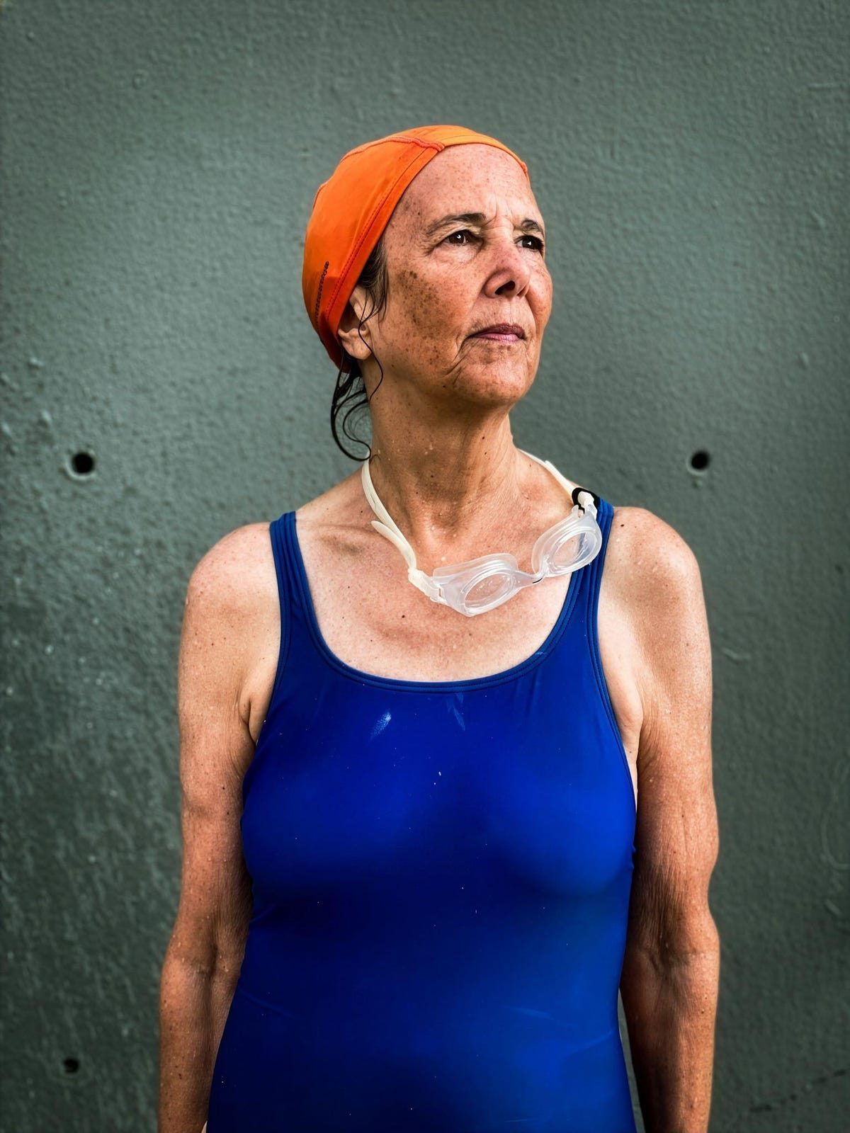 A woman in a blue swimsuit and orange swim cap looks to the side. Swim goggles hand from her neck.