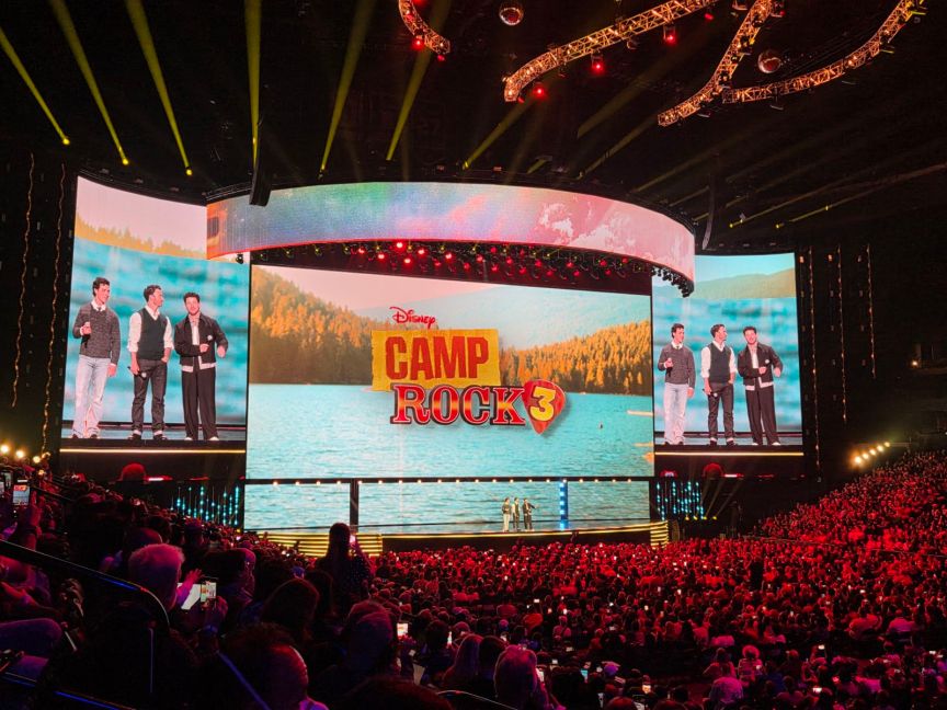 Three men stand on stage in front of a screen with the words "Disney Camp Rock 3" in front of a crowd of people.