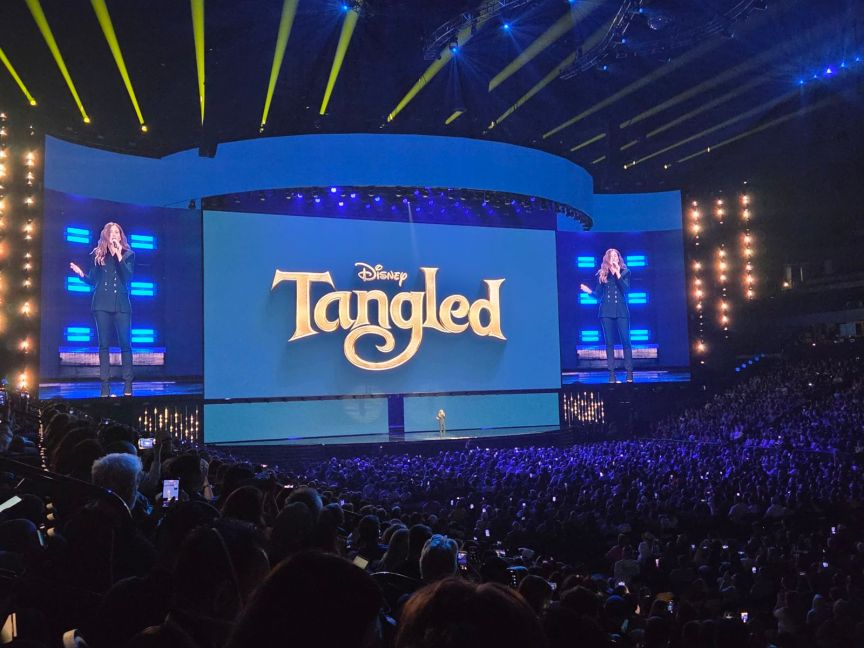 A woman stands on stage in front of a screen with the words "Disney Tangled" in front of a crowd of people.