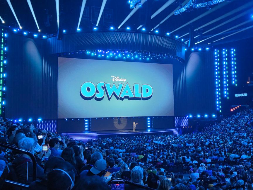 A man stands on stage in front of a screen with the words "Disney Oswald" in front of a crowd of people.