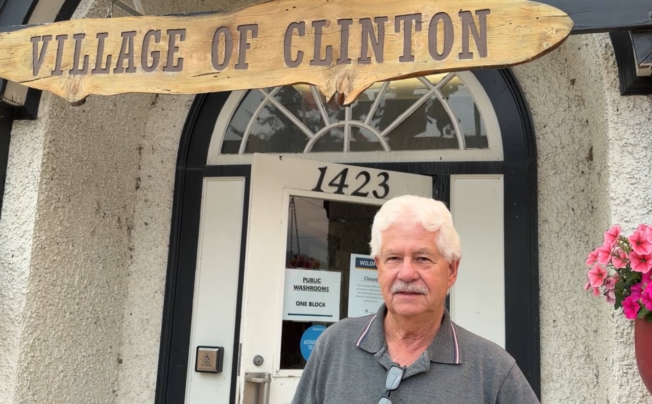 A man with grey hair and a moustache stands in front of a building with a wooden sign that says "Village of Clinton."
