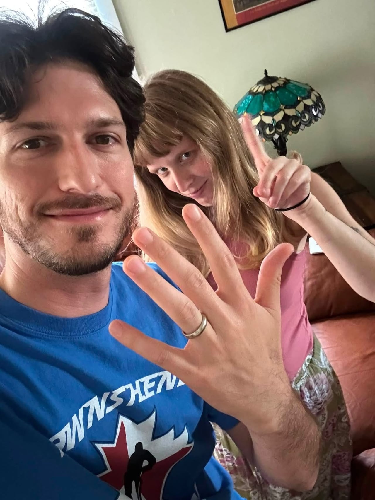 A person holds up his hand to show a wedding band. A woman in the background wags her finger.