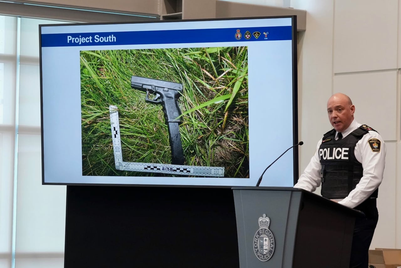 Police officer stands at a podium discussing investigation with a photo of a gun on a TV screen beside him.