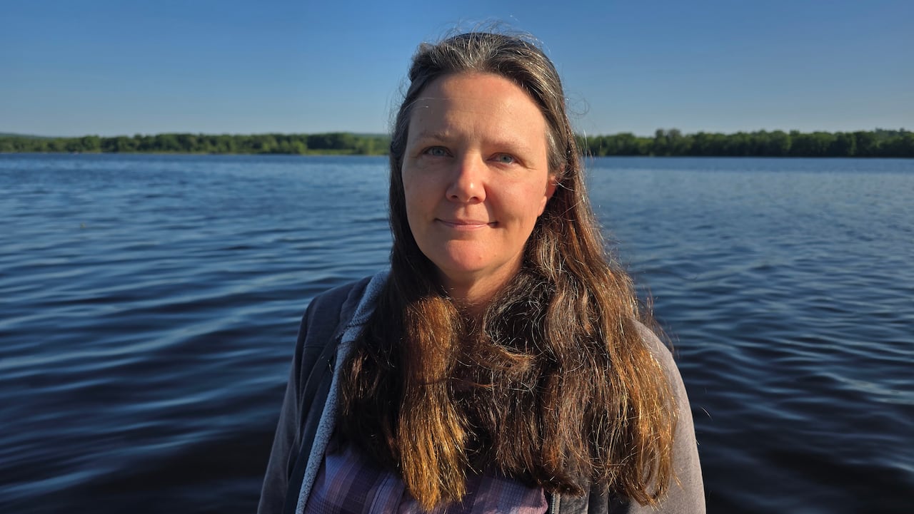 A woman with brown hair stands with a river behind her as she faces the camera.