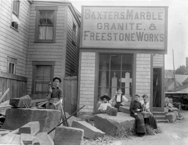 A black and white photo of a old building with a man hammer and chiseling stones out front. Four woman and a small dog sit nearby. A large sign on the top of the building reads "Baxters Marble Granite, & Freestone Works."