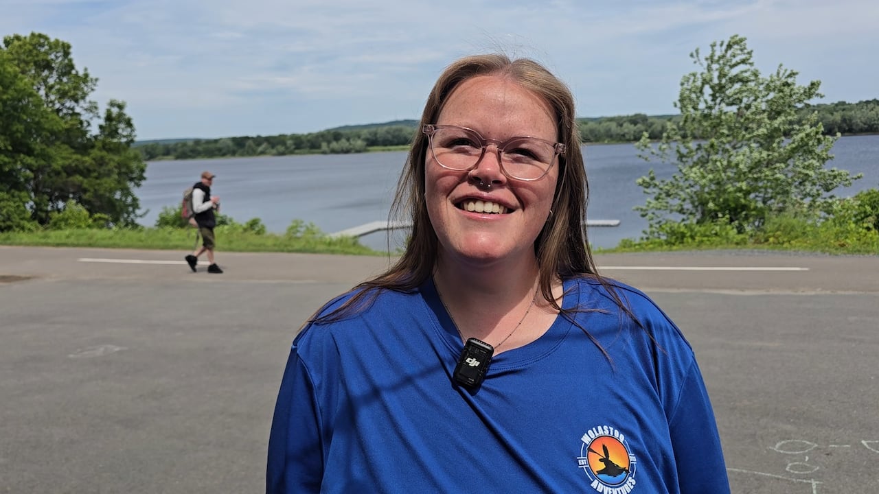 A woman with light brown hair wearing clear-rimmed glasses and a blue t-shirt with a nose and septum piercing smiles at the camera with a walking trail and river behind her.