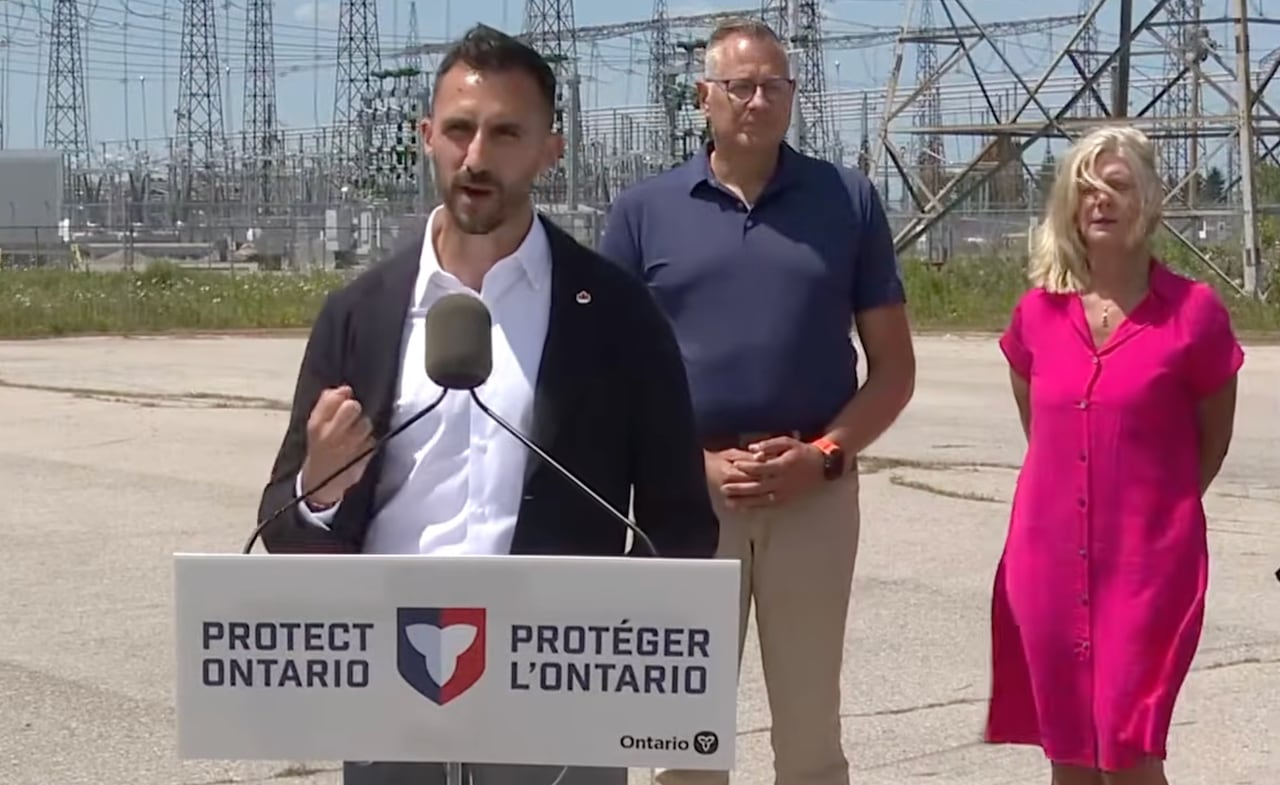 Ontario Energy and Mines Minister Stephen Lecce at a lecturn is flanked by two other people with transmission lines in the background.