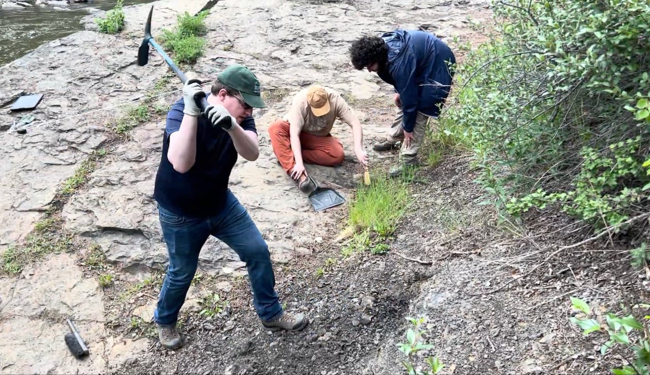 A man in a green hat and jeans swings a pick axe against a rocky riverbank while two others behind him crouch inspecting the ground.