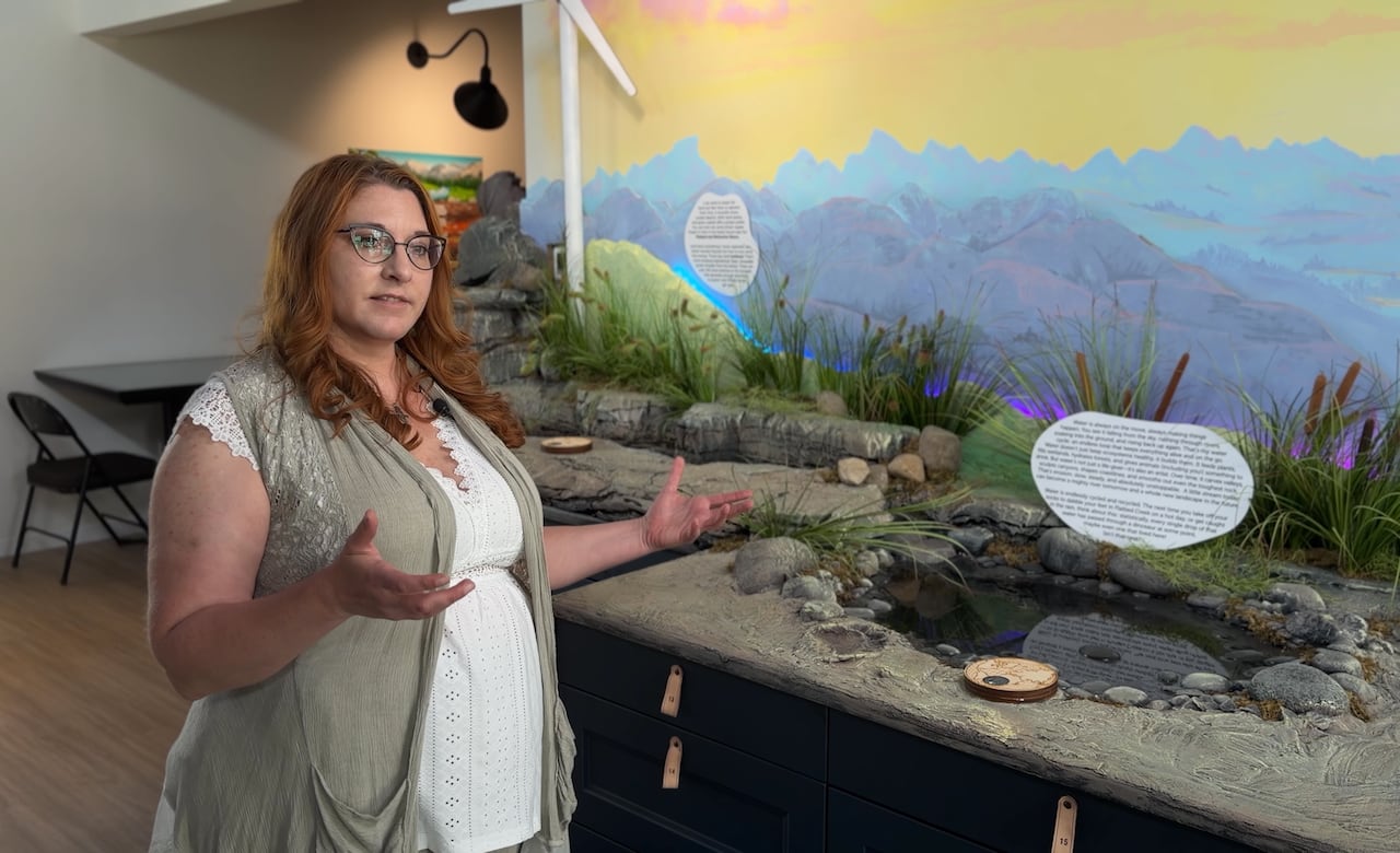 A professionally dressed woman with red hair and glasses motions to a museum display of a prehistoric pond with fossil tracks set against a mountain landscape.