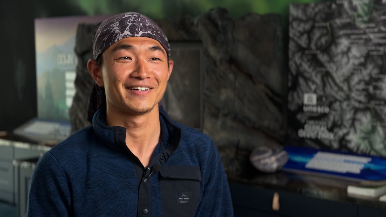 A young and smiling Asian man wearing a dark blue pullover and purple patterned bandana.