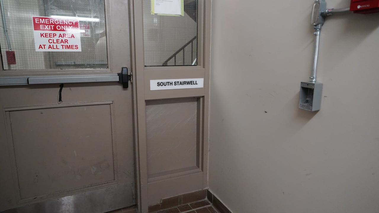 A beige hallway is shown with a door leading to a stair way next to an empty, metal electrical box on the wall.