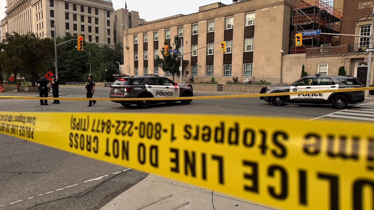Several police officers stand outside the US Consulate building in downtown Toronto near dawn on a summer morning. The building is taped off with police tape. A police cruiser is parked on the street with lights flashing.