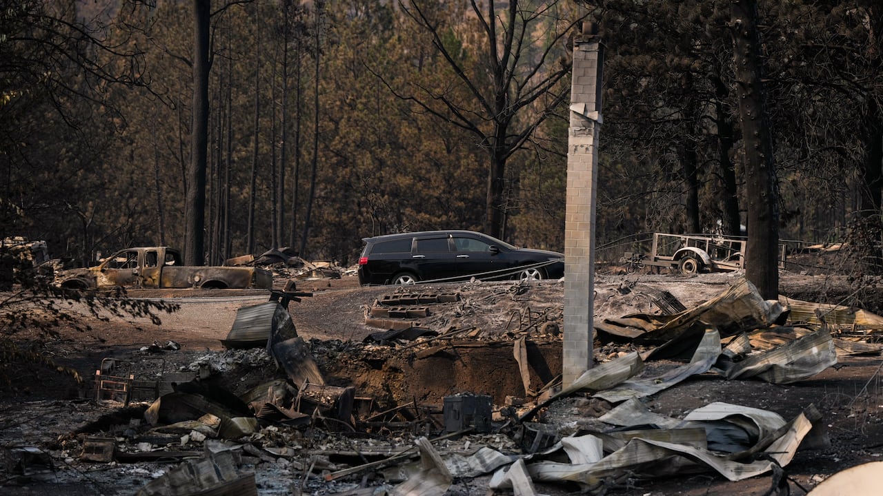 In the foreground, you can see burned-out wreckage of a home with only a chimney still standing. In the background is a forest, with some trees blackened and a charred pickup truck.