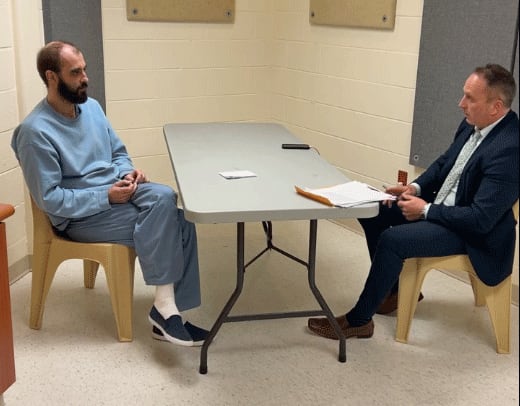 A man wearing blue jail-issued top and pants sitting at a table across from a man wearing a suit.