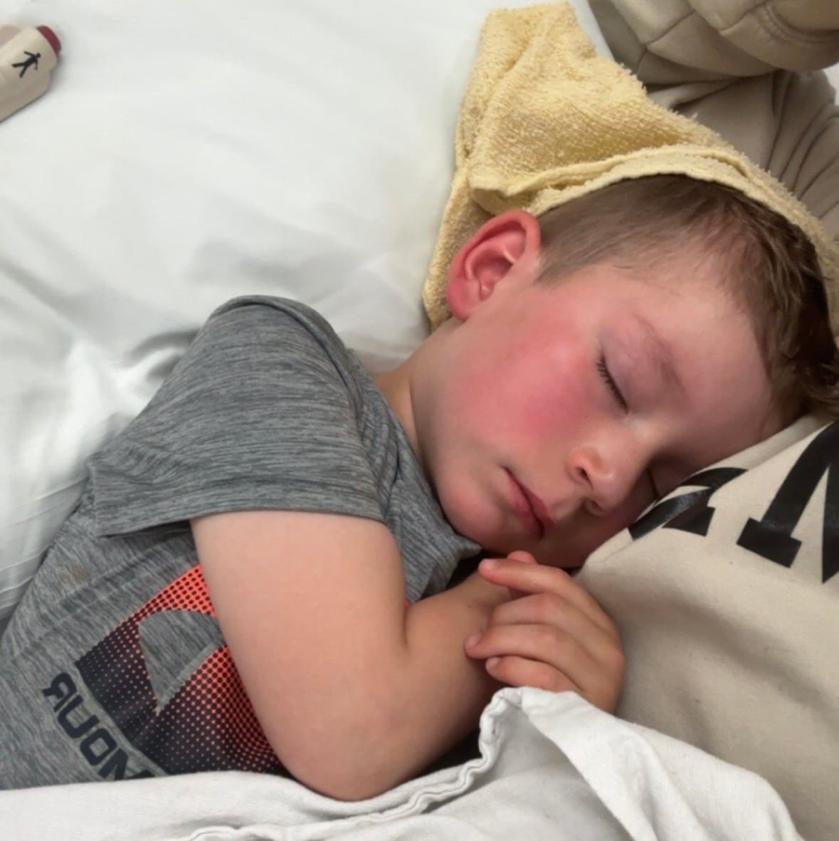 A boy with rosy cheeks from a fever sleeps in his mom's arms in a hospital bed.