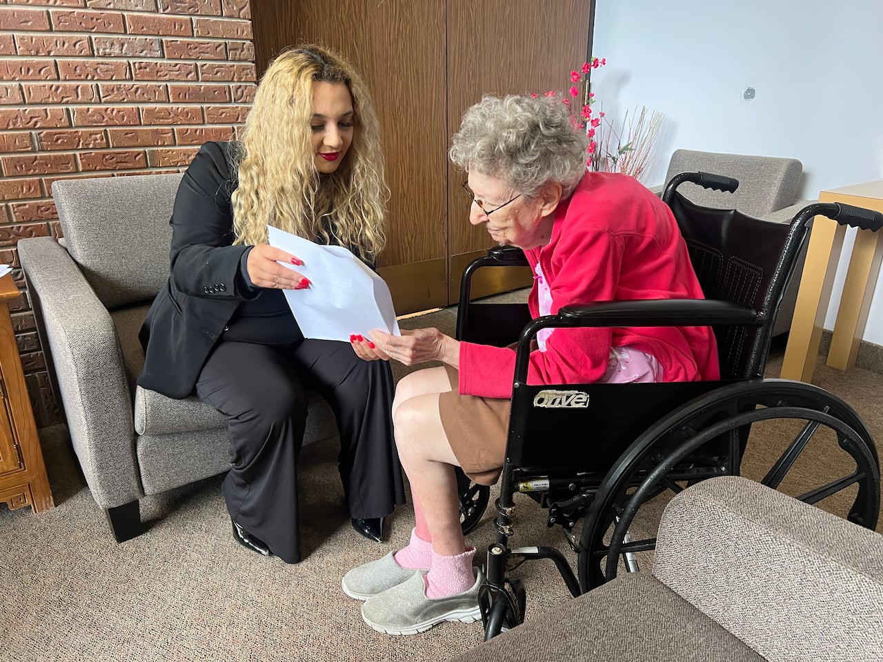 A woman in black business attire shows a woman in a wheelchair a printed document