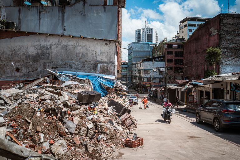 Motorists and pedestrians pass by the wreckage of a collapsed building on a street in Buenaventura. [Euan Wallace / Al Jazeera]