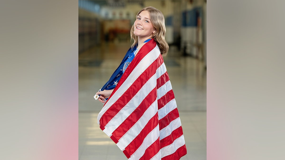 A teenage girl in a blue Northwest No. 30 lacrosse uniform holds an American flag outstretched behind her in a school hallway.