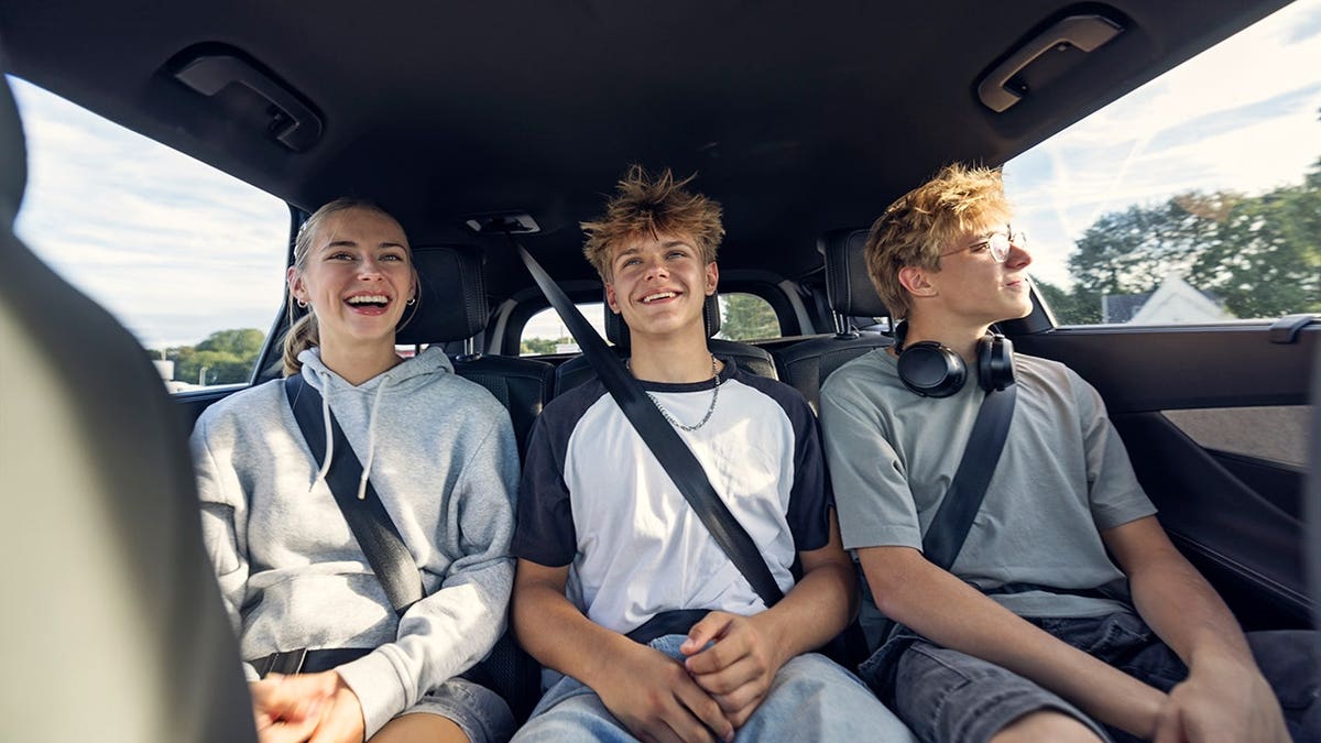 Three teenagers sit in the backseat of a car being driven through a town.