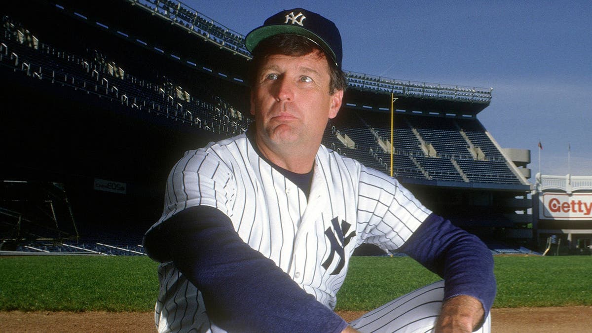Tommy John in Yankees uniform looks on from the dugout at Yankee Stadium.