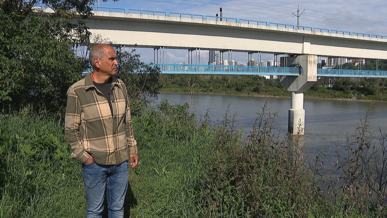 Man wearing a plaid jacket and jeans looking out at the North Saskatchewan River. Behind him is the Edmonton LRT bridge.