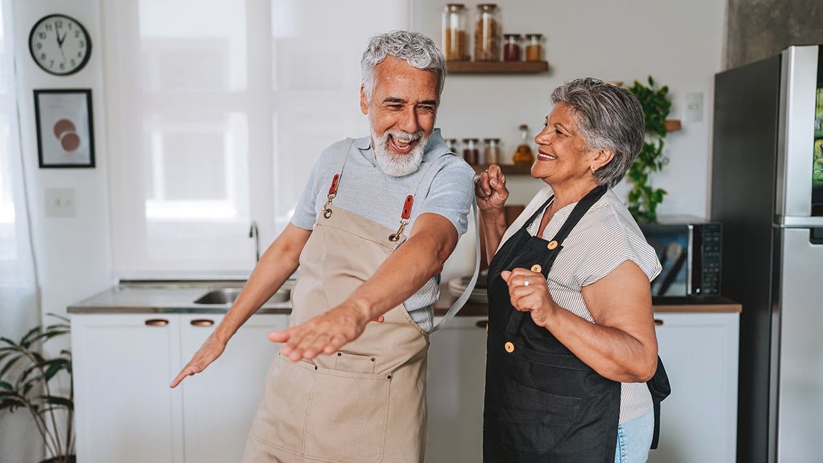 Senior couple dances and smiles while wearing aprons in a kitchen.
