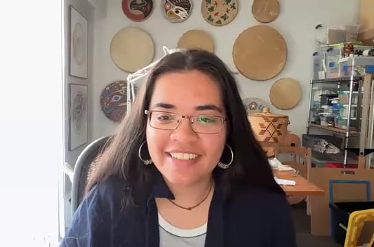A smiling woman with long black hair and glasses, seen on a Zoom call, circular pieces of woven artwork hanging on the wall behind her