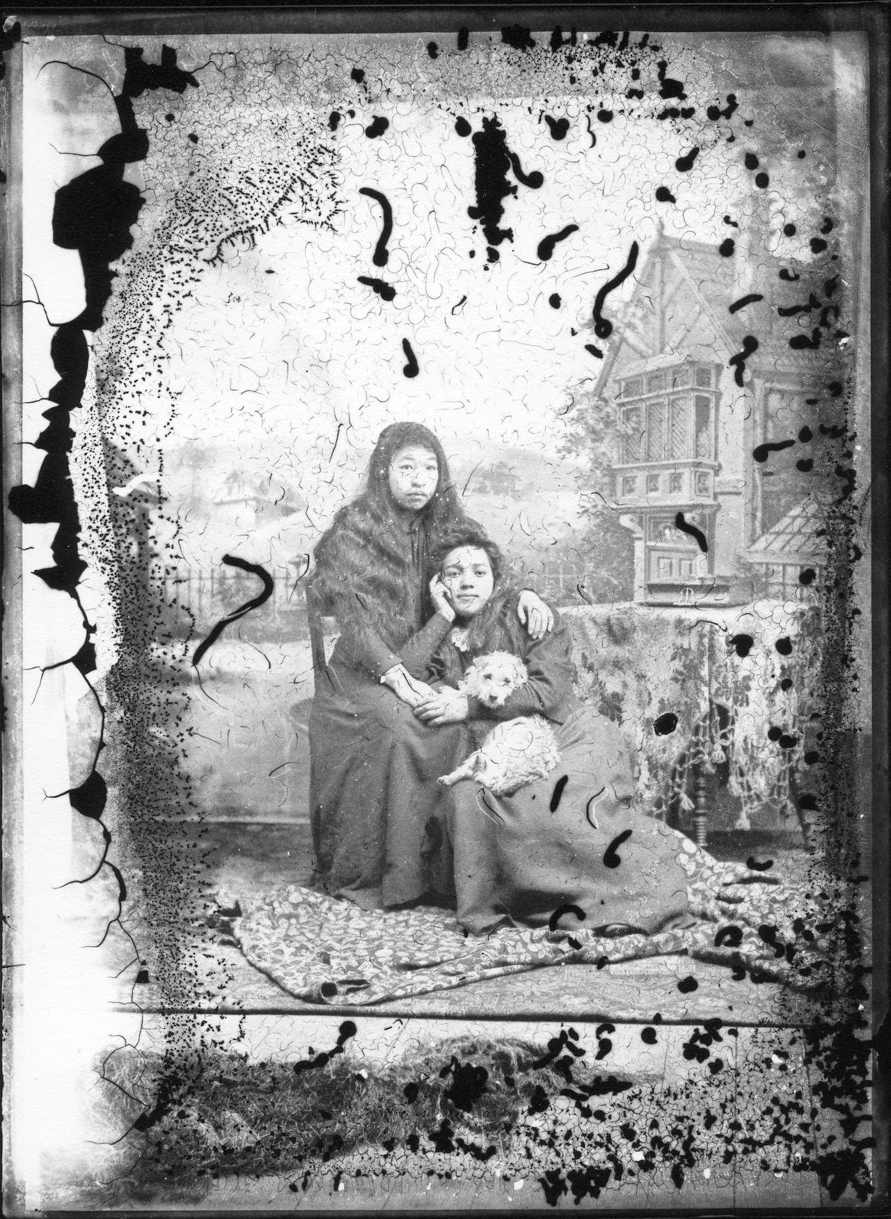 A black and white photo, wit black spots of damage, showing two girls posing together in a chair with a small fluffy dog.