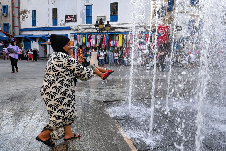 A woman carries a child as they cool off by water jets from a fountain at the entrance to the Medina in Tunis on August 24, 2026, amid a heatwave with temperatures exceeding 48°C and daily water and electricity cuts [Fethi Belaid/AFP]